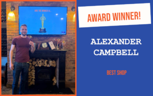 The winner of the evening's question at the April Cambridge networking event, standing in front of a fireplace, holding the trophy. Behind them is a screen displaying a trophy.